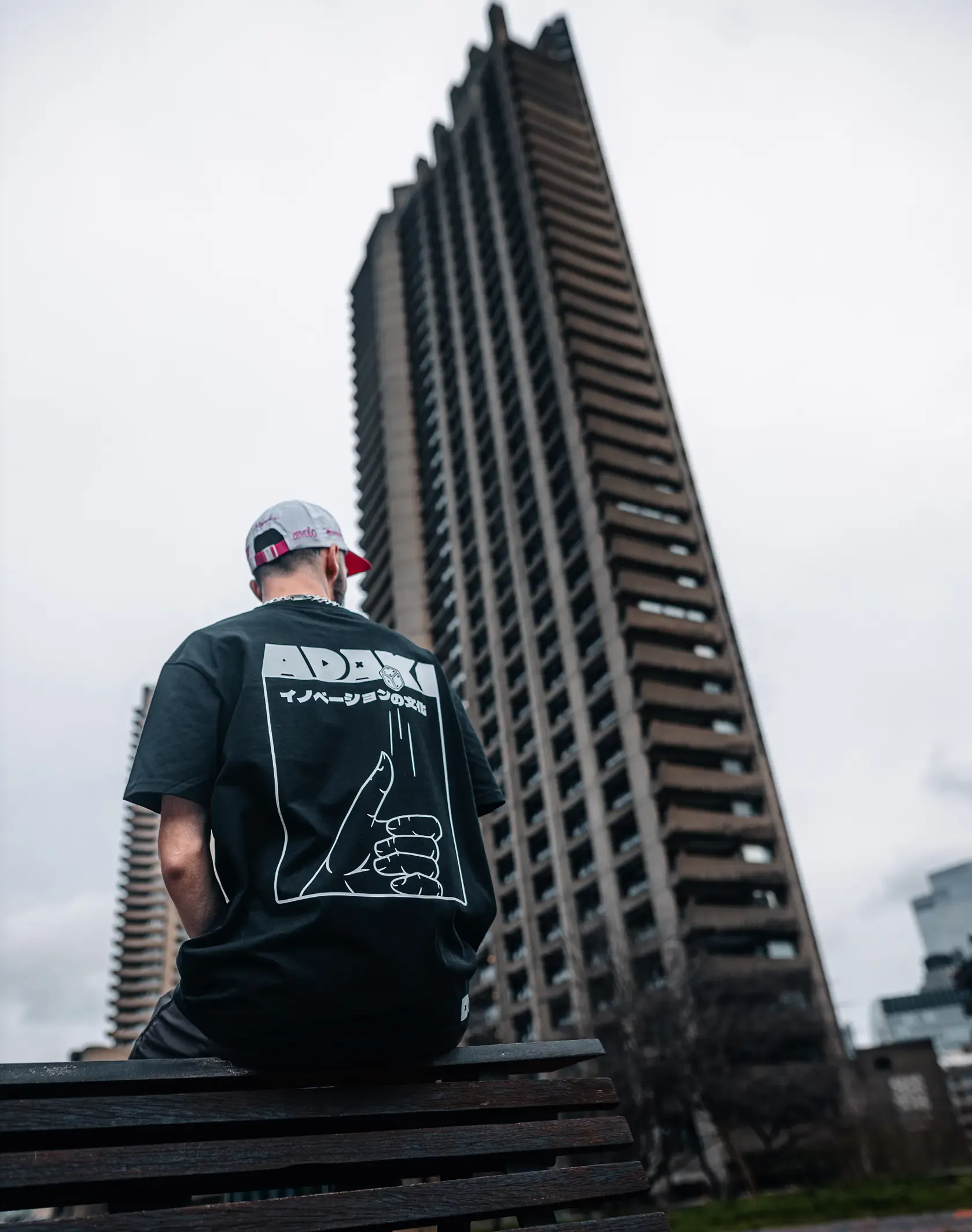 A photograph of a man sat on a bench at Barbican in London, showing the back of the Adaki T-shirt
