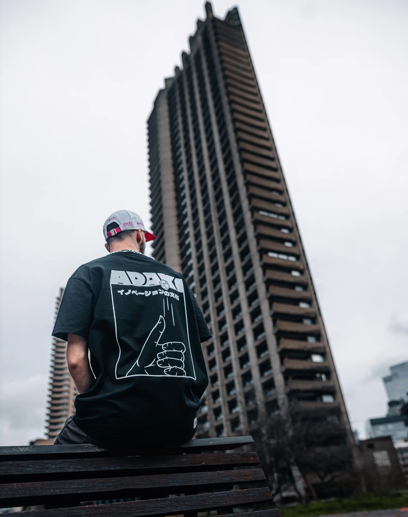 A photograph of a man sat on a bench at Barbican in London, showing the back of the Adaki T-shirt